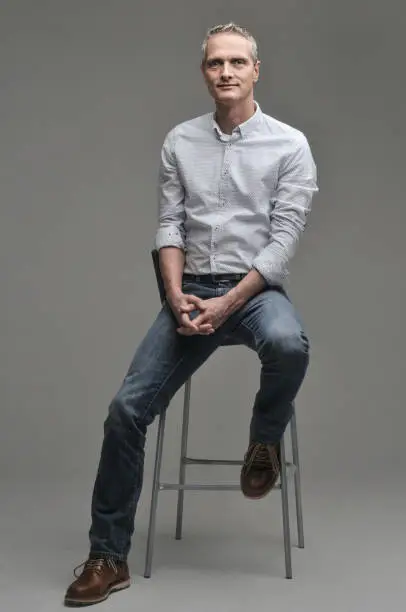 Photo of Man in a shirt and jeans is sitting on a chair.
Gray background.
