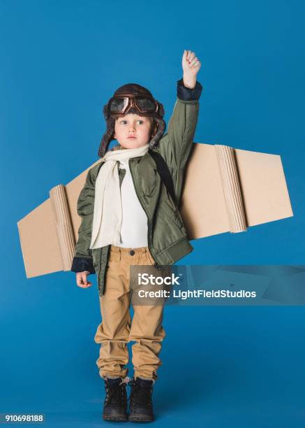 Cute Little Boy In Pilot Costume With Paper Plane Wing Isolated On Blue Stock Photo - Download Image Now
