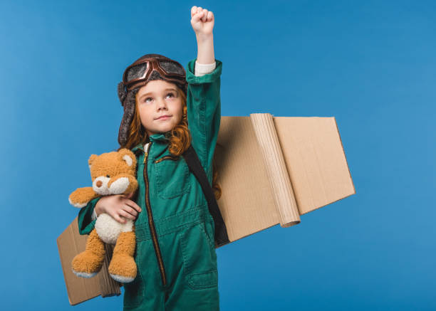 portrait of cute child in pilot costume with teddy bear and handmade paper plane wings isolated on blue portrait of cute child in pilot costume with teddy bear and handmade paper plane wings isolated on blue alternative pose photos stock pictures, royalty-free photos & images
