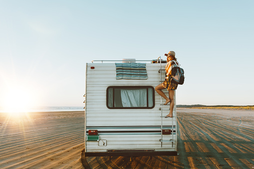Charming young woman with nice smile with hat, sunglasses, backpack climing on recreational vehicle on the ocean beach at sunset.