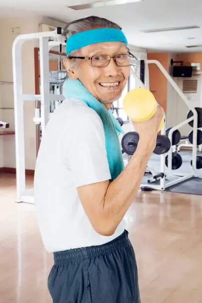 Side view of elderly man smiling at the camera while exercising with a dumbbell in the fitness center