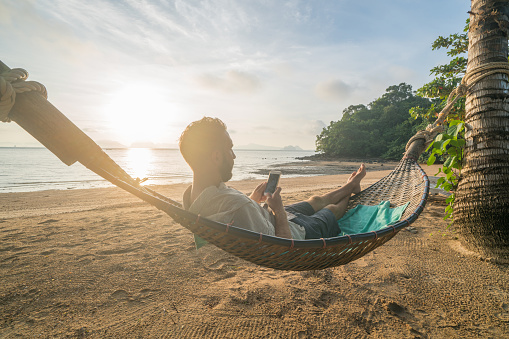 Young man relaxing on hammock at sunrise using mobile phone, set on a tropical beach in the southern Islands of Thailand. Beautiful light and sunbeam.