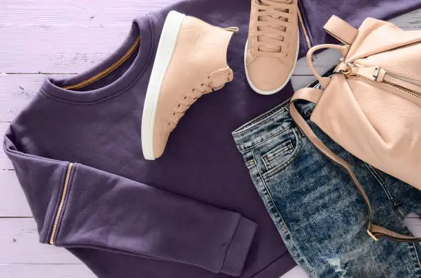 Photo of Womens clothing, accessories, footwear (violet sweatshirt, acid washed jeans,  leather backpack and sneakers) on purple wooden background. Outfit for teens. Top view, flat lay. Trendy colors