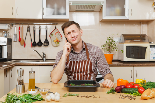 Perturbed stress young man in apron sitting at table with vegetables, talking on mobile phone, cooking at home preparing meat stake from pork, beef or lamb, in light kitchen with wooden surface