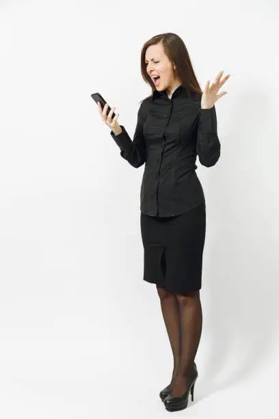 Full length portrait of angry caucasian young brown-hair business woman in black classic shirt and skirt talking, screaming on mobile phone isolated on white background. Copy space for advertisement