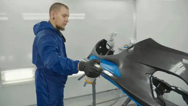 Serviceman preparing a car bodykit for painting in a workshop. Vehicle bumper before paint in spray booth.