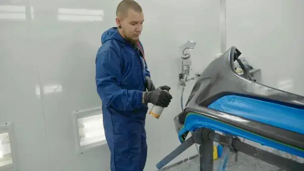 Serviceman preparing a car bodykit for painting in a workshop. Vehicle bumper before paint in spray booth.