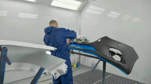 Serviceman preparing a car bodykit for painting in a workshop. Vehicle bumper before paint in spray booth.