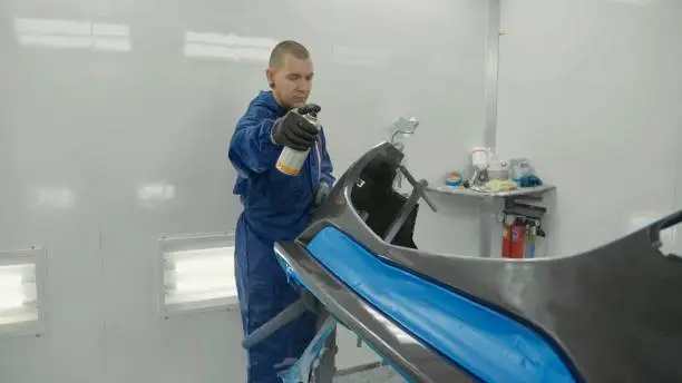 Serviceman preparing a car bodykit for painting in a workshop. Vehicle bumper before paint in spray booth.