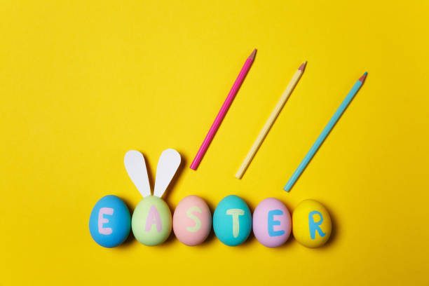 istock Row of colorful pastel monophonic painted Easter eggs with inscription Easter, fun bunny ears on egg, three pencils on yellow background. Copy space for advertisement. With place for text. Top view. 910802484
