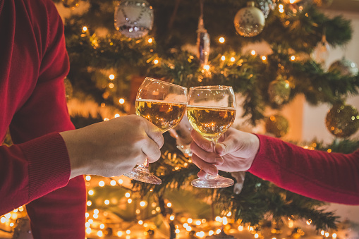 Close up of male and female hands cheering and holding wine glasses in front of decorated Christmas tree. New year eve celebration.