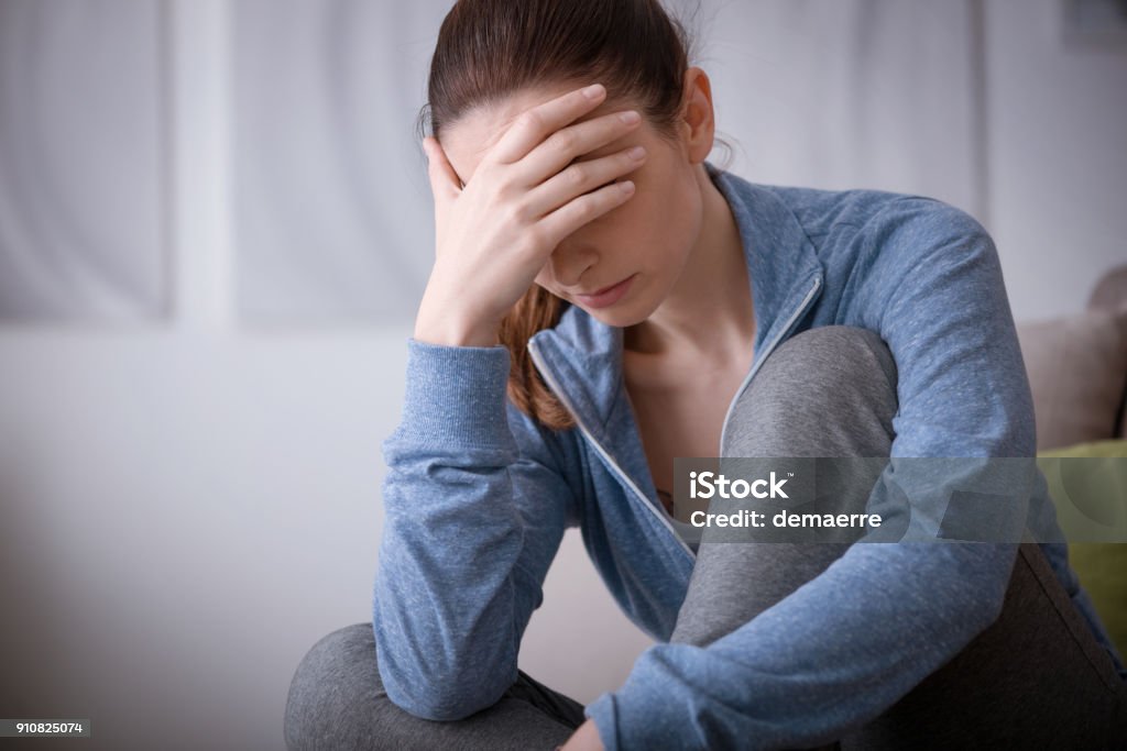 Depressed woman at home Sad depressed woman at home sitting on the couch, looking down and touching her forehead, loneliness and pain concept Headache Stock Photo