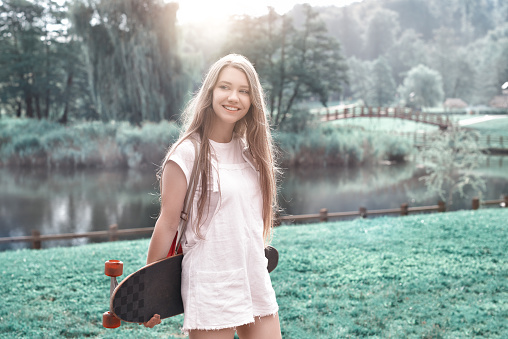 smiling young woman in the park holding her skateboard and enjoying summer day.