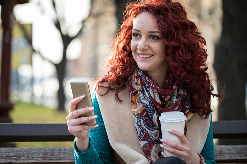 Cute redhead with smart phone and coffee to go sitting on the bench in park