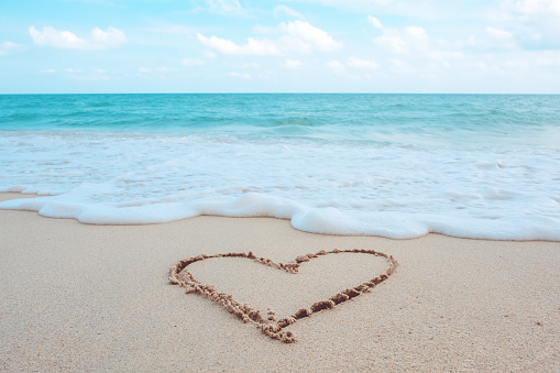 The hand writing heart shaped on the beach by the sea with white waves and blue sky background