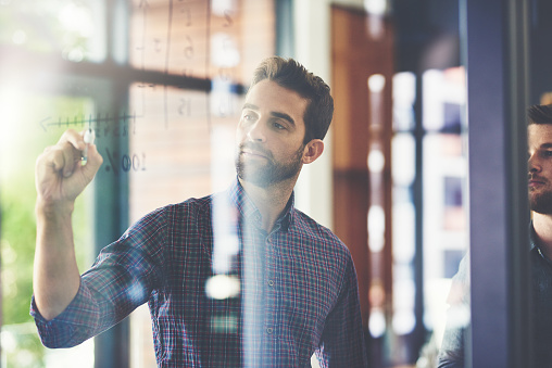 Shot of two young businessmen brainstorming on a glass wall in an office