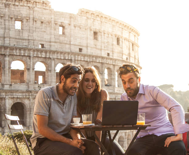 istock Three happy friends using laptop having video call sitting at bar restaurant table in front of colosseum in rome at sunset 910879846