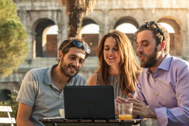 istock Three happy friends using laptop having video call sitting at bar restaurant table in front of colosseum in rome at sunset 910879888
