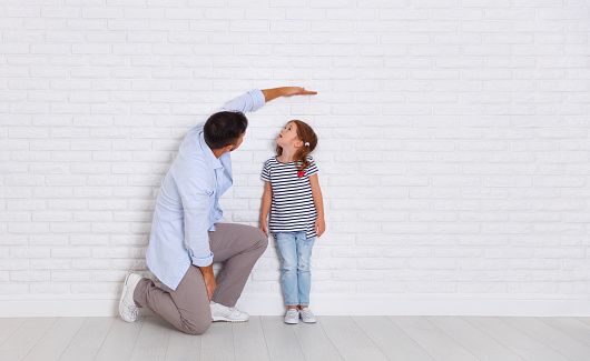 concept of the family. the father measures growth of child to a daughter near an empty wall