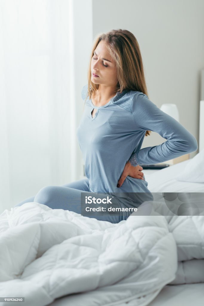 Woman with back pain Young attractive woman with back pain waking up and sitting on her bed, she is touching her back Backache Stock Photo