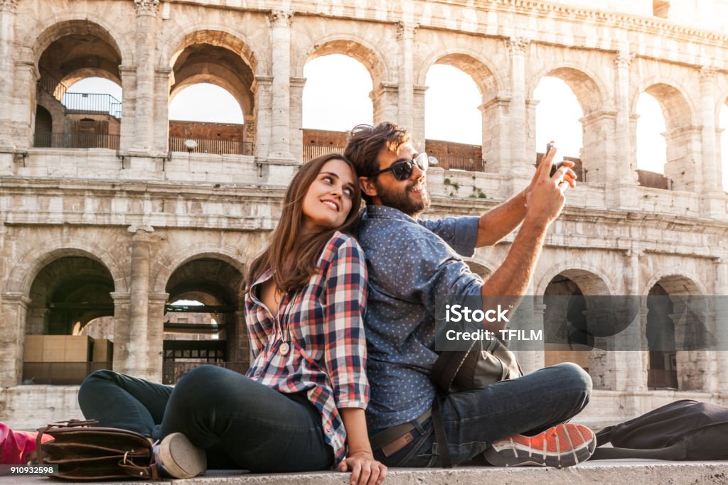 Happy couple sitting in front of colosseum in rome taking selfie pictures with smartphone camera. Sunset with lens flare Happy couple sitting in front of colosseum in rome taking selfie pictures with smartphone camera. Sunset with lens flare. Travel Stock Photo