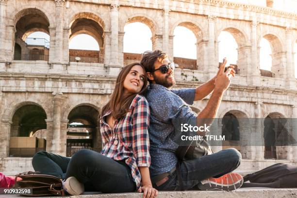 Happy Couple Sitting In Front Of Colosseum In Rome Taking Selfie Pictures With Smartphone Camera Sunset With Lens Flare Stock Photo - Download Image Now