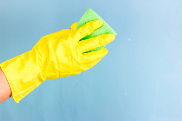 istock Washing windows and glass. The worker in a yellow glove can stack a sponge with foam on a blue background 910987634