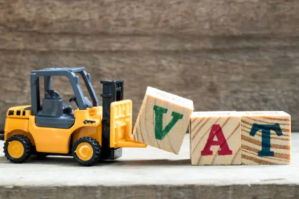 Toy plastic forklift hold block V to compose and fulfill wording VAT on wood background