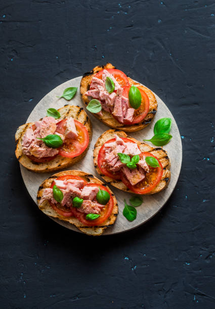 sanduíches com tomate, manjericão e atum na tábua de madeira sobre um fundo escuro, vista superior. pequeno-almoço saudável, tapas, snack - bruschetta - fotografias e filmes do acervo