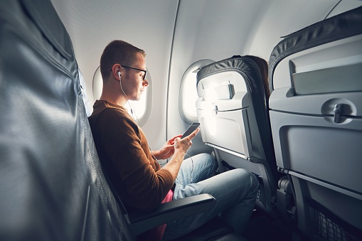 Connection in the airplane. Young man (traveler) using smart phone during flight and listening music.