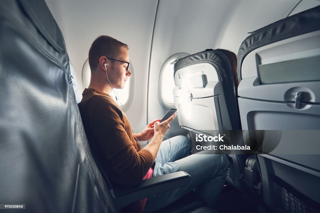 Connection in the airplane Connection in the airplane. Young man (traveler) using smart phone during flight and listening music. Airplane Stock Photo