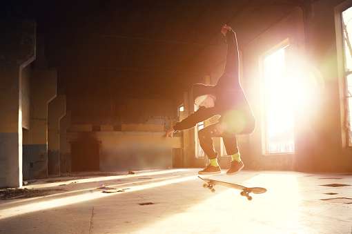 A young skater in a white hat and a black sweatshirt does a trick with a skate jump in an abandoned building in the backlight of the setting sun