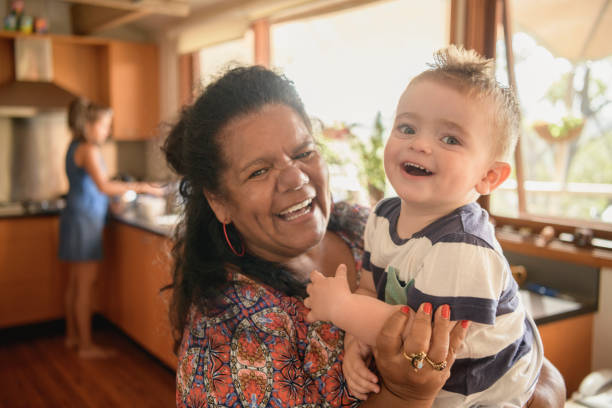 Portrait of Aboriginal grandmother holding baby grandson Young boy and mature woman smiling towards camera, mother in background doing dishes australian culture stock pictures, royalty-free photos & images