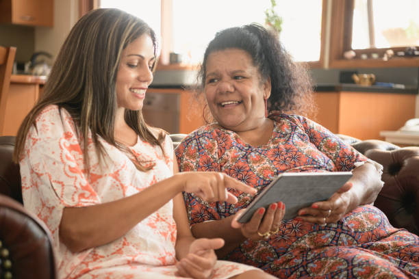 Two women using digital tablet smiling and laughing Aboriginal young woman showing her mother how to use tablet, pointing at the screen australian culture stock pictures, royalty-free photos & images
