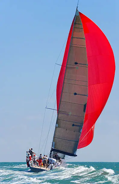 A fully crewed racing yacht with a red spinnaker catching the wind and leaving a big wake