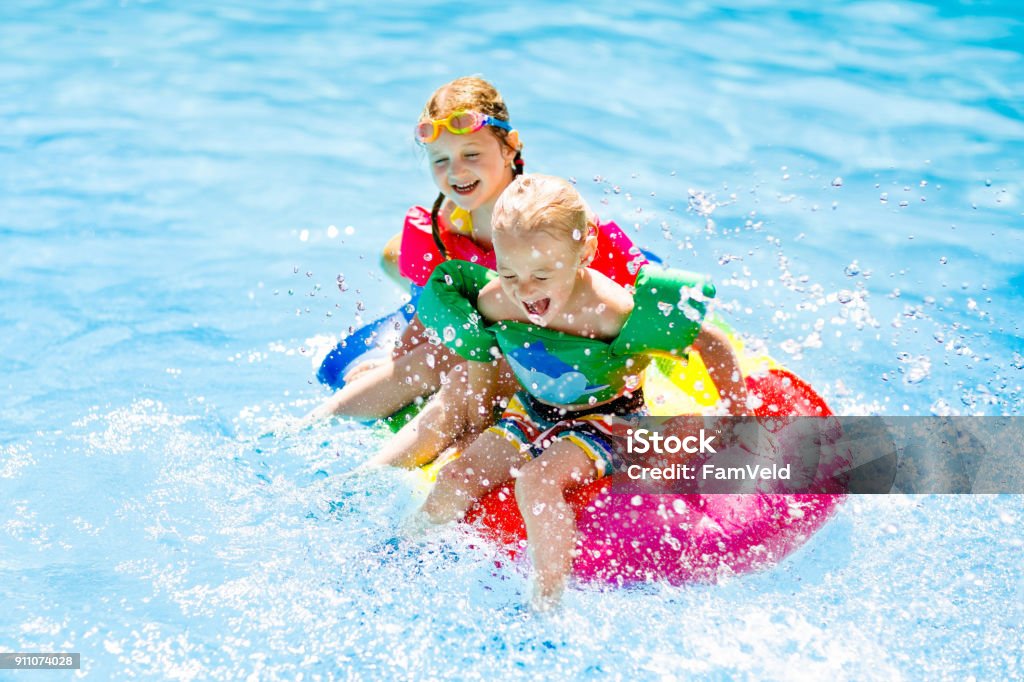 Kids on inflatable float in swimming pool. Boy and girl on inflatable ice cream float in outdoor swimming pool of tropical resort. Summer vacation with kids. Swim aids and wear for children. Water toys. Little child floating on colorful raft. Swimming Pool Stock Photo