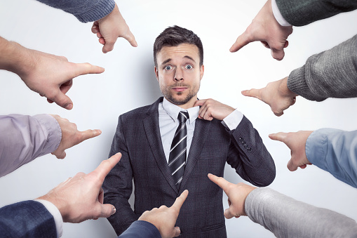 Businessman stretching his collar while many index fingers are pointing at him.
