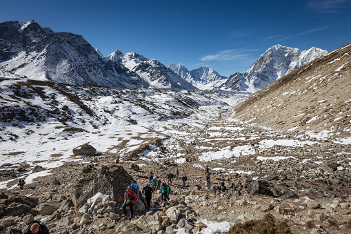 a large group of hikers and walkers making their way up the valley close to Mount Everest base camp, Nepal, with beautiful snow capped mountains in the distance and a blue sky, and very rocky terrain