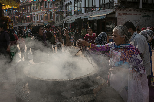 a Nepalese woman standing beside a large smoking pot in Kathmandu, with dramatic sunlit backlit lighting, and crowds of people and tourists in the background