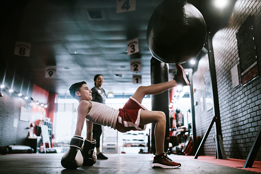 A youth trains hard at a martial arts training gym, learning kickboxing.  Good exercise, self discipline and self defense. His coach is visible in the background behind him.