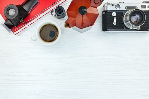retro camera, roll films, lens, notebook and grunge red coffee pot on white wooden background