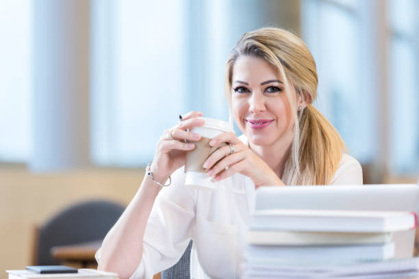 Beautiful mid adult female college student smiles while studying in the campus library. She is drinking a cup of coffee.
