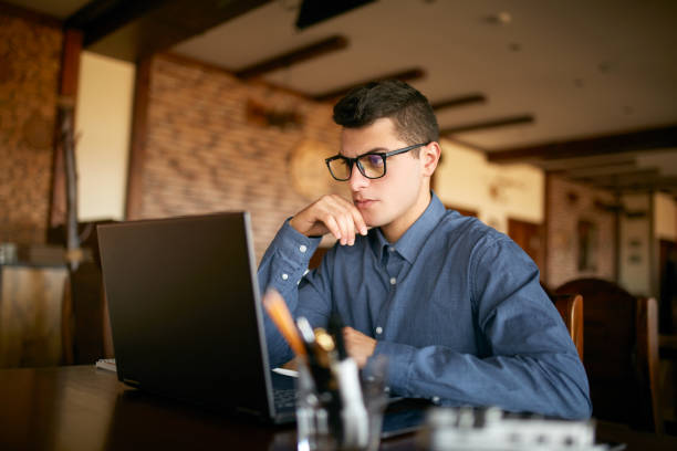 istock Thoughtful young caucasian businessman in glasses working on laptop computer. Pensive attractive hipster freelancer thinks over the solution of the problem with computer in cafe. Business concept 911336732