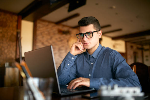 istock Thoughtful young caucasian businessman in glasses working on laptop computer. Pensive attractive hipster freelancer thinks over the solution of the problem with computer in cafe. Business concept 911336762