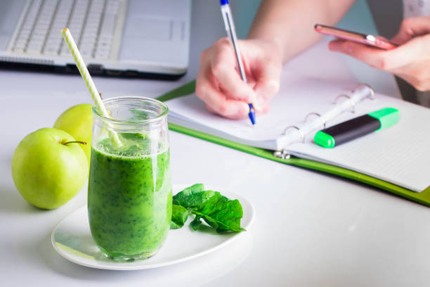 여자의 오른손 및 왼손에 노트북에 쓰기 전화 모바일 스마트 폰, 노트북 근처를 보유 하고있다. 녹색 시금치와 사과에서 건강 음료 스무디 - dieting juice green freshness 뉴스 사진 이미지