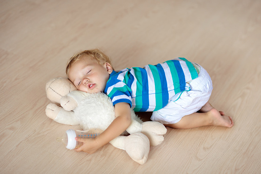 Baby sleeping on wooden floor with stuffed toy sheep and milk bottle. Funny tired little boy falling asleep crawling on hardwood floor at home. Sleepy infant drinking formula on soft cushion.