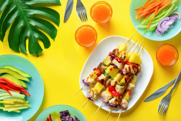 Summer simple recipe for grilling, hawaiian chicken kabobs served with freshly diced vegetables and some orange drink in glasses, view from above