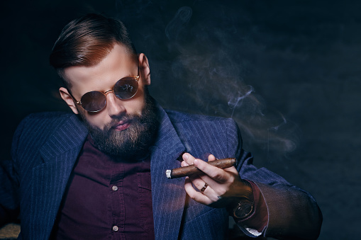 Bearded modern man in sunglasses smoking a cigar.