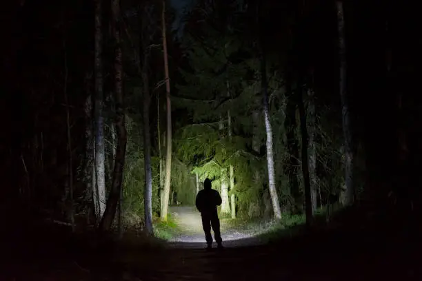 Photo of Man standing in dark forest at night with flashlight and hoodie on head
