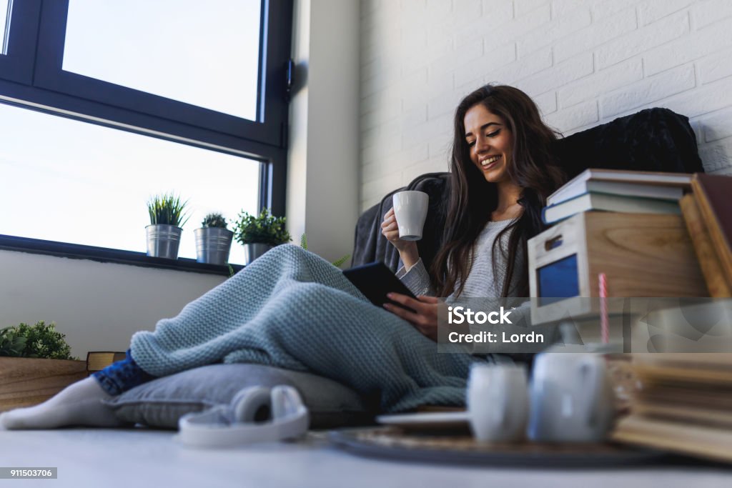 Beautiful young smiling woman reading eBook reader at home Beautiful young woman reading eBook reader at home surrounded by books Book Stock Photo